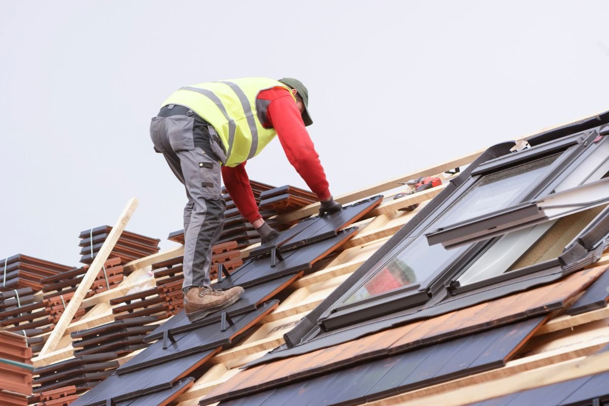 A man installs a skylight on a metal roof, standing near a window, with tools and materials around him.